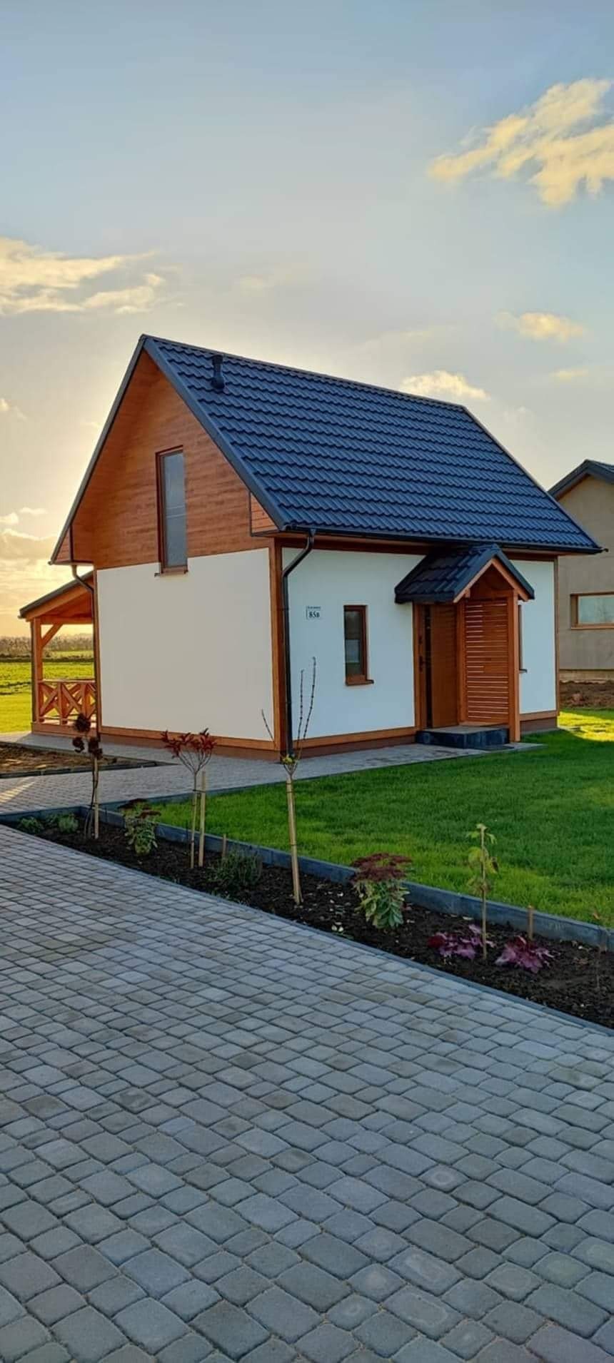 Gotowy, parterowy dom z drewnianą elewacją, ciemnym dachem, małym gankiem i zadbanym trawnikiem oraz kostką brukową na podjeździe, oświetlony zachodzącym słońcem.