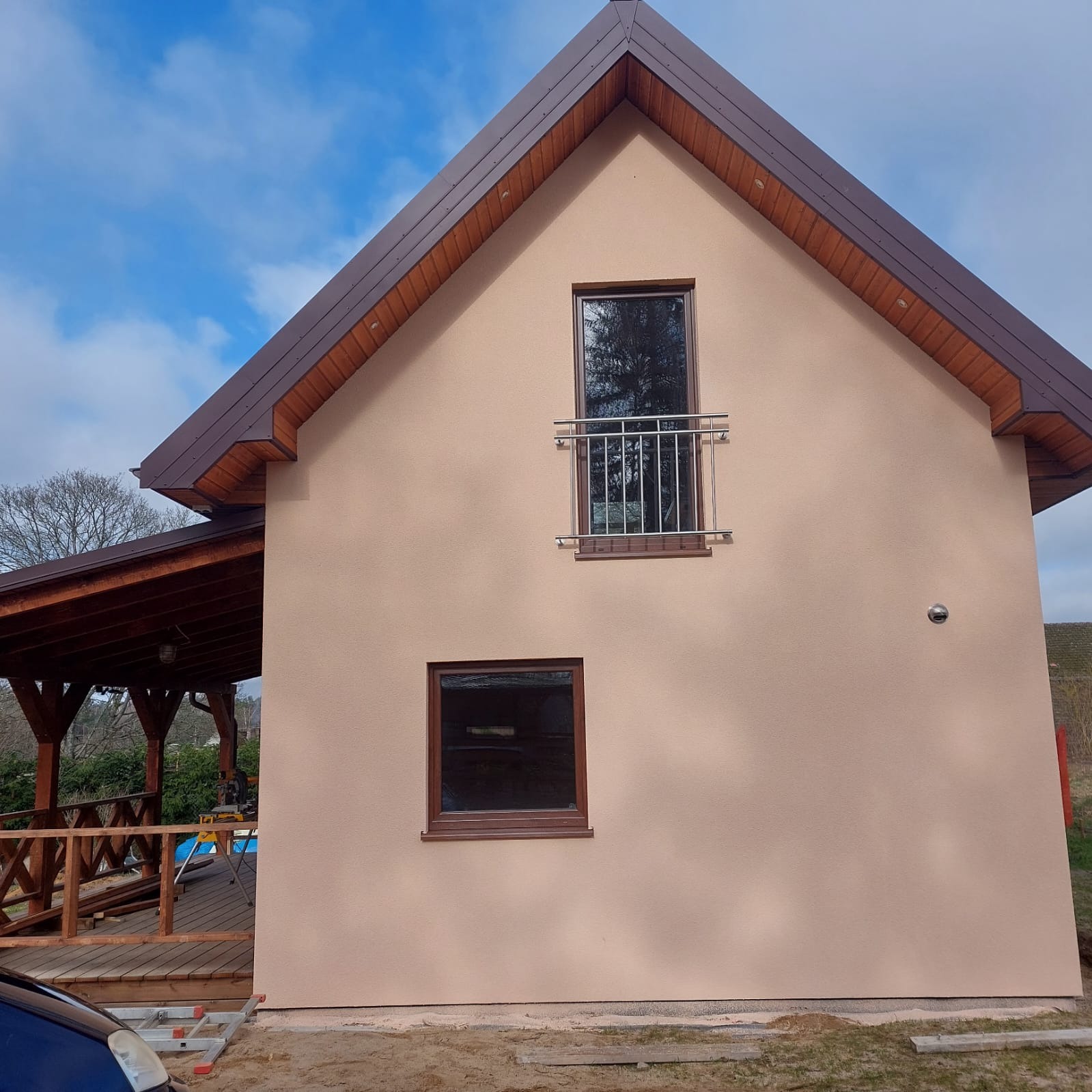Elewacja domu jednorodzinnego z beżowym tynkiem, brązowym dachem i drewnianą altaną z boku, widok z poziomu gruntu.