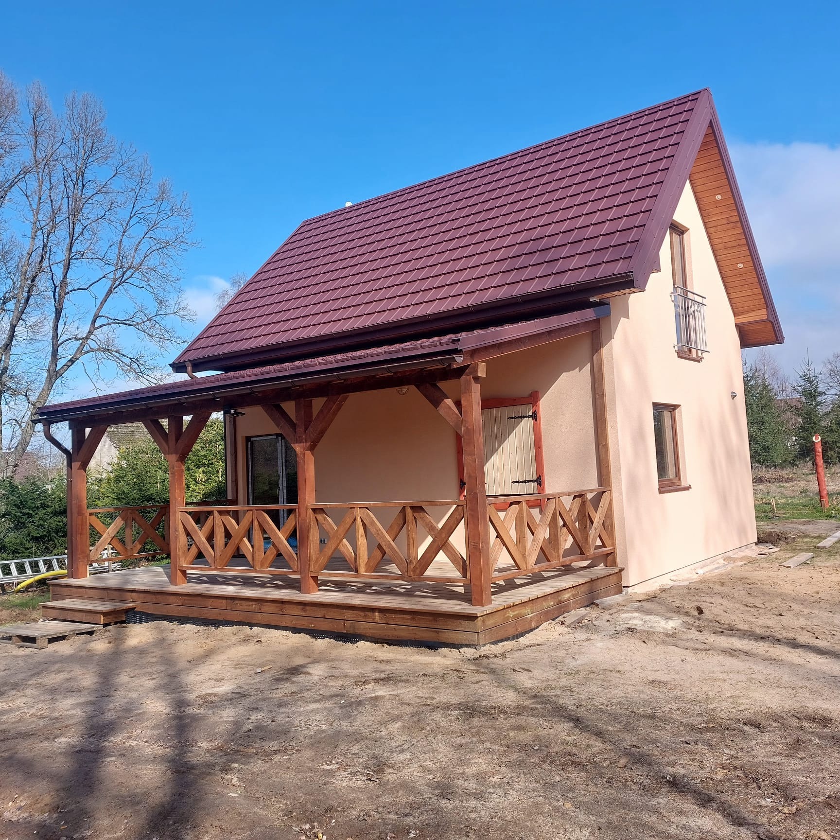 Niewielki, parterowy dom z poddaszem, zadaszonym tarasem z drewnianą balustradą w kształcie krzyży, brązowy dach z blachodachówki, elewacja w kolorze beżowym.