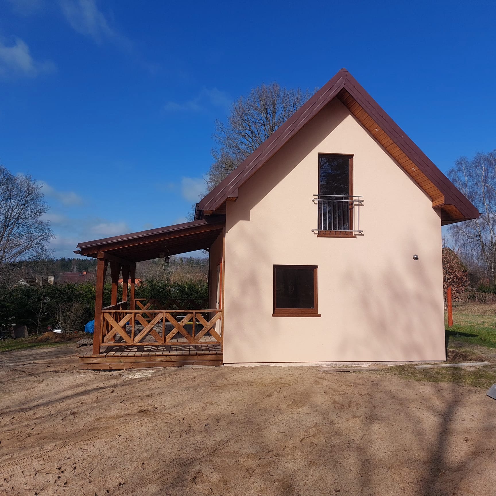 Nieduży, jasny dom z brązowym dachem i drewnianą werandą, widok z zewnątrz w słoneczny dzień.