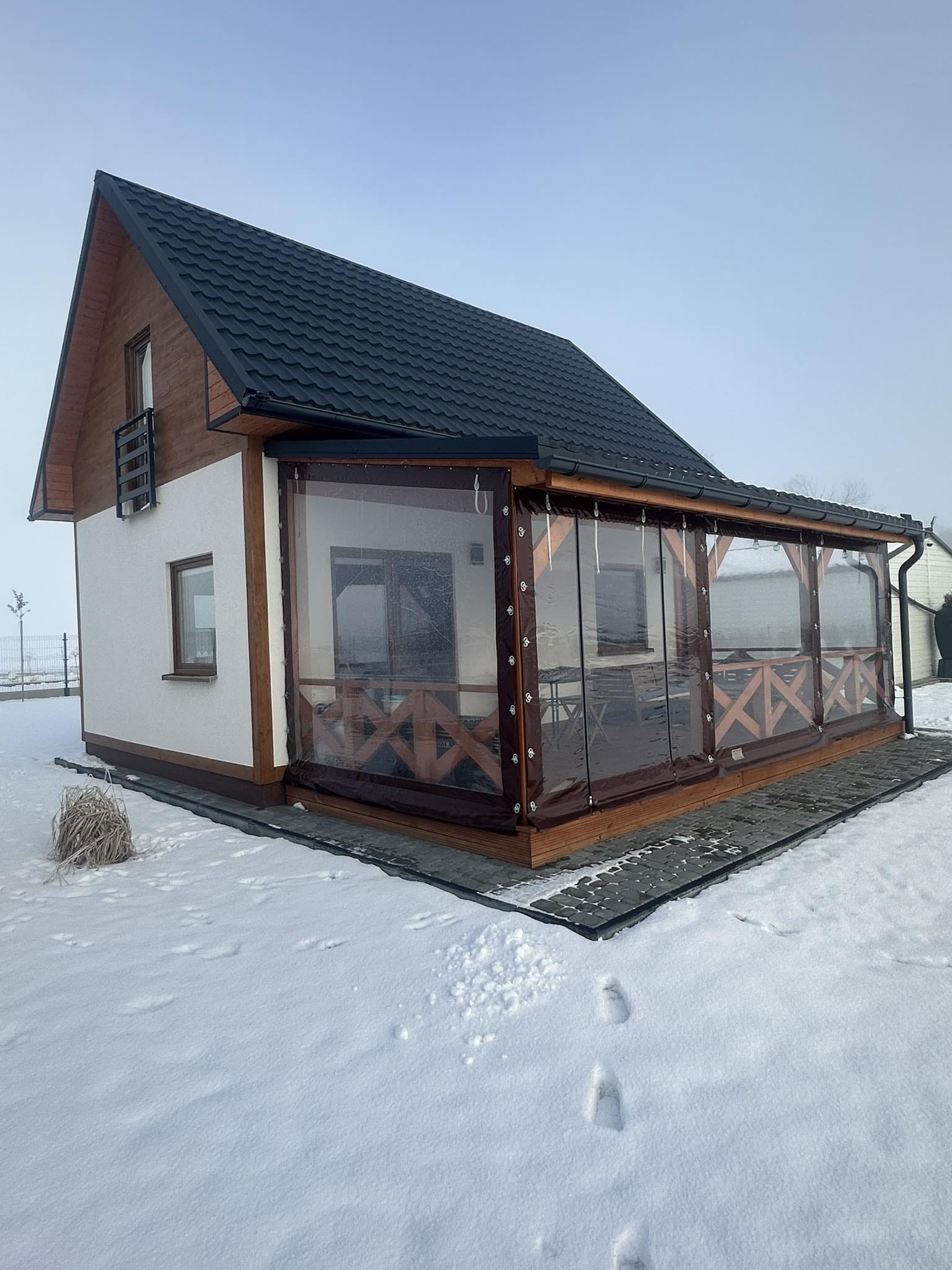 Niewielki dom z dobudowaną oszkloną werandą, pokryty czarną dachówką, otoczony śniegiem, widoczne ślady stóp na śniegu.
