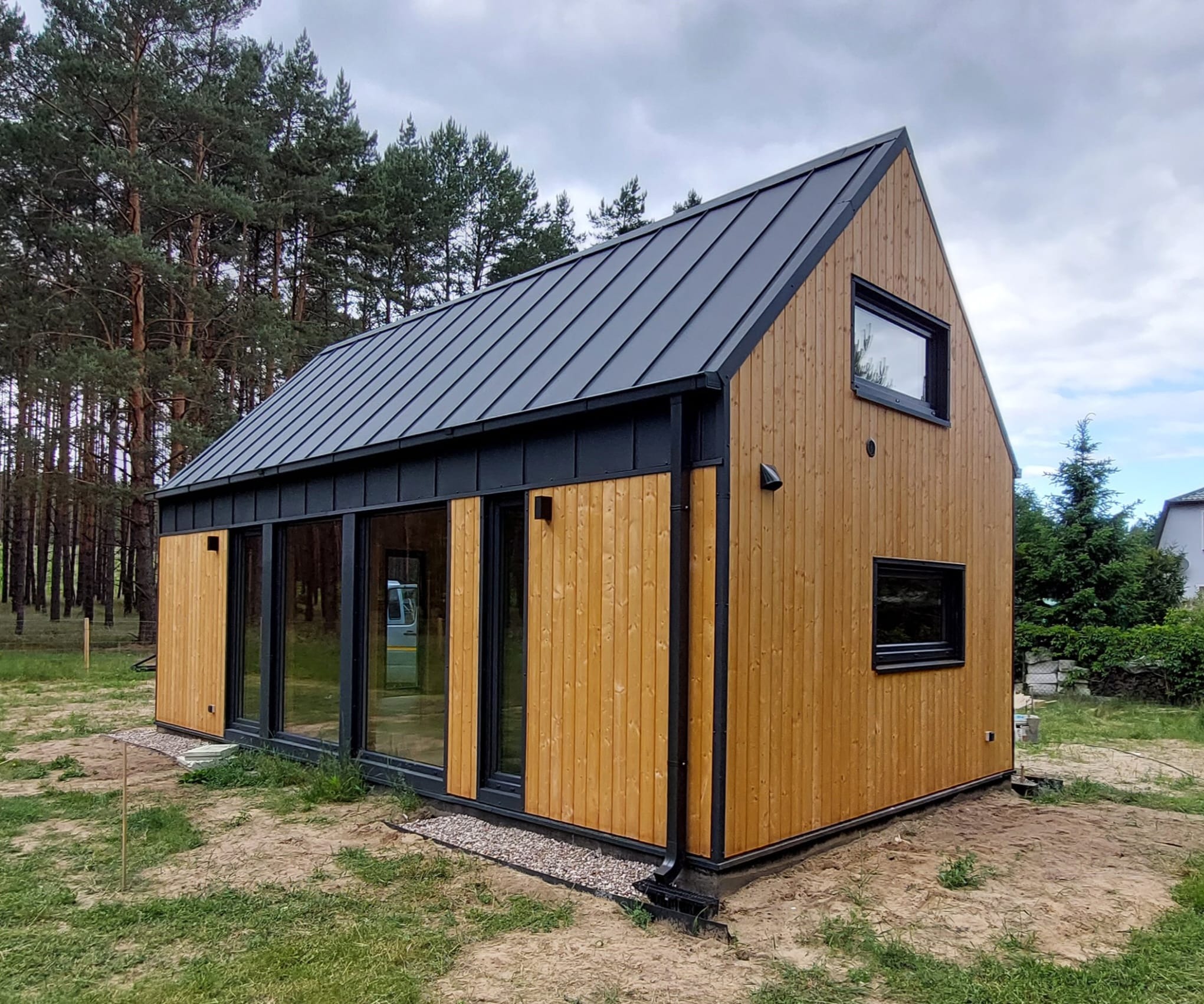 Nowoczesny dom z drewna o prostej bryle, z grafitową blachą na dachu, dużymi oknami i elewacją w naturalnym kolorze drewna, otoczony zielenią.