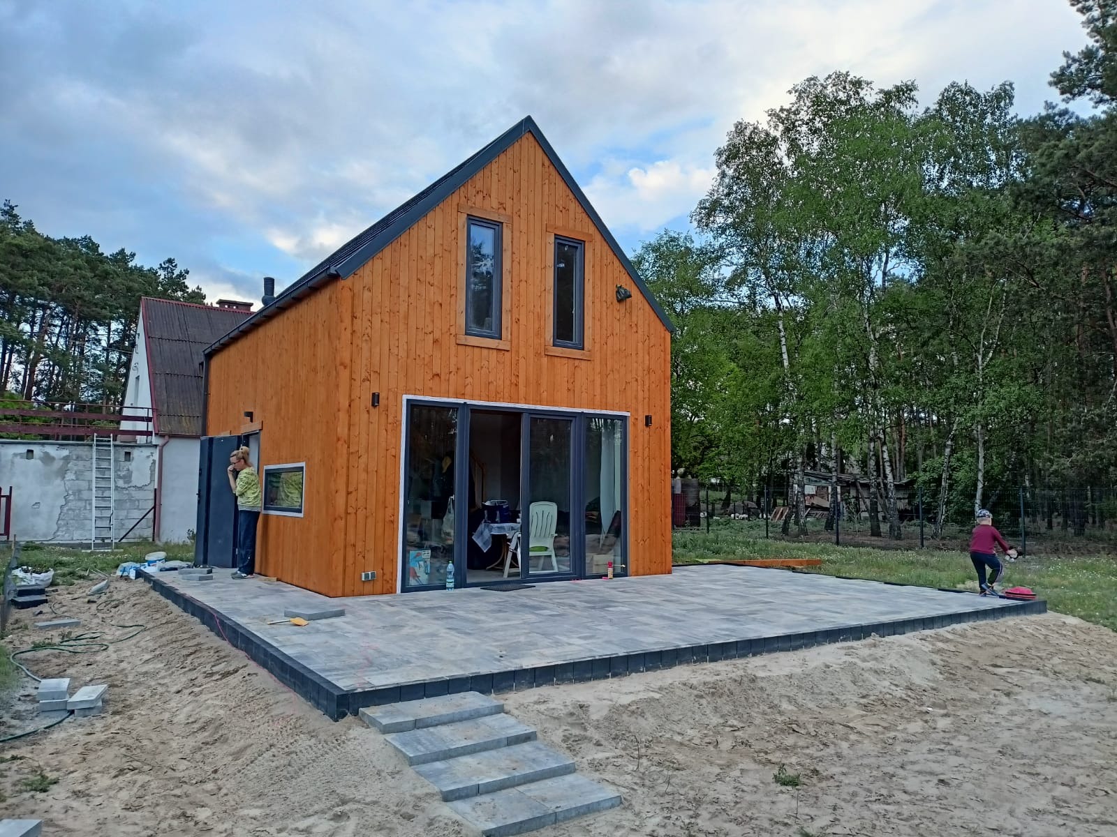 Nowoczesny dom szkieletowy z drewnianą elewacją i tarasem z kostki brukowej w trakcie budowy, widoczne osoby i narzędzia.