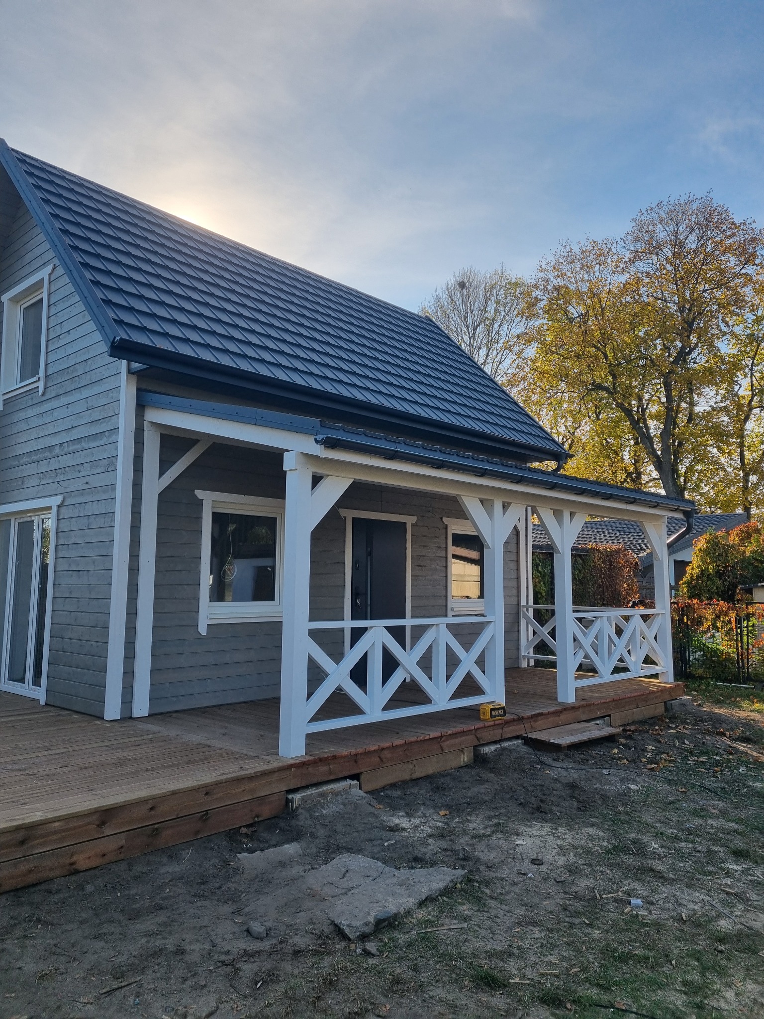 Nowoczesny dom z szarego drewna z ciemnym dachem i białym zadaszonym gankiem z drewnianą podłogą, widoczne elementy krajobrazu w tle.