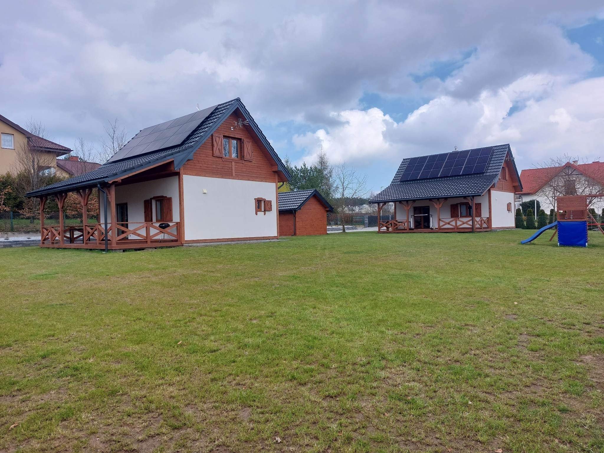 Dwa parterowe domy z panelami słonecznymi na dachu, drewnianymi werandami i białymi ścianami, otoczone rozległym trawnikiem i innymi budynkami w tle.