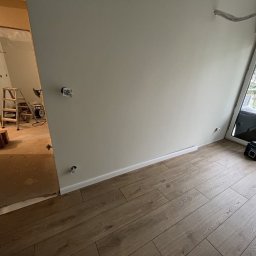 Usługi Remontowo-Budowlane Koza Marek - Wnętrze w trakcie prac wykończeniowych: świeżo pomalowana ściana w odcieniu szarości, drewnopodobna podłoga, widoczna drabina i narzędzia w sąsiednim pomieszczeniu, okno balkonowe z widokiem...