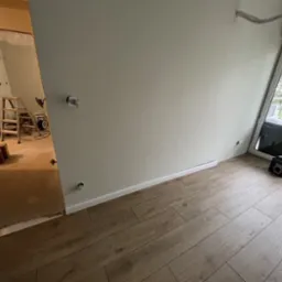 Wnętrze w trakcie prac wykończeniowych: świeżo pomalowana ściana w odcieniu szarości, drewnopodobna podłoga, widoczna drabina i narzędzia w sąsiednim pomieszczeniu, okno balkonowe z widokiem...