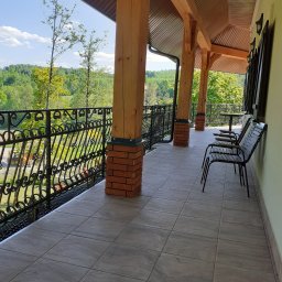 Usługi Remontowo-Budowlane Koza Marek - Zadaszony balkon z drewnianymi filarami na ceglanej podstawie, ozdobną metalową balustradą i szarymi płytkami podłogowymi, z widokiem na zielony krajobraz w tle.