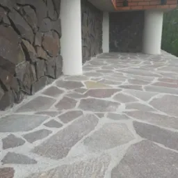 Kamienna elewacja budynku z nieregularną okładziną i kolumnami, widoczna kamienna posadzka balkonu z szarych płyt o nieregularnych kształtach.