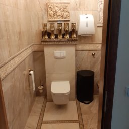 Usługi Remontowo-Budowlane Koza Marek - Elegancka łazienka w beżowych marmurkowych płytkach z dekoracyjnymi elementami: płaskorzeźby nad toaletą i złotymi świecznikami, czarny kosz na śmieci, widok zza otwartych drzwi.