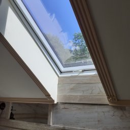 Usługi Remontowo-Budowlane Koza Marek - Łazienka z oknem dachowym, widok na błękitne niebo i korony drzew, marmurowe płytki ścienne z dekoracyjnym wzorem, złota bateria umywalkowa, białe kubki na szczoteczki do zębów.