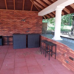 Usługi Remontowo-Budowlane Koza Marek - Zadaszona przestrzeń z ceglaną ścianą, kolumną i rattanowymi meblami, wykończona czerwoną kostką brukową, z widocznymi opadłymi liśćmi.