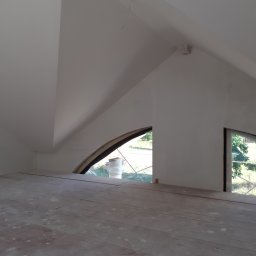 Usługi Remontowo-Budowlane Koza Marek - Poddasze w trakcie prac wykończeniowych, widoczne okna o nietypowych kształtach, surowe deski podłogowe pokryte pyłem, białe ściany i skosy. Widok z poziomu podłogi.