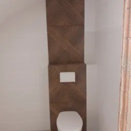 Nowoczesna toaleta podwieszana z drewnopodobną zabudową stelaża i geometrycznym wzorem, białe płytki na ścianie z delikatną falą, szara podłoga, surowy mur w narożniku.