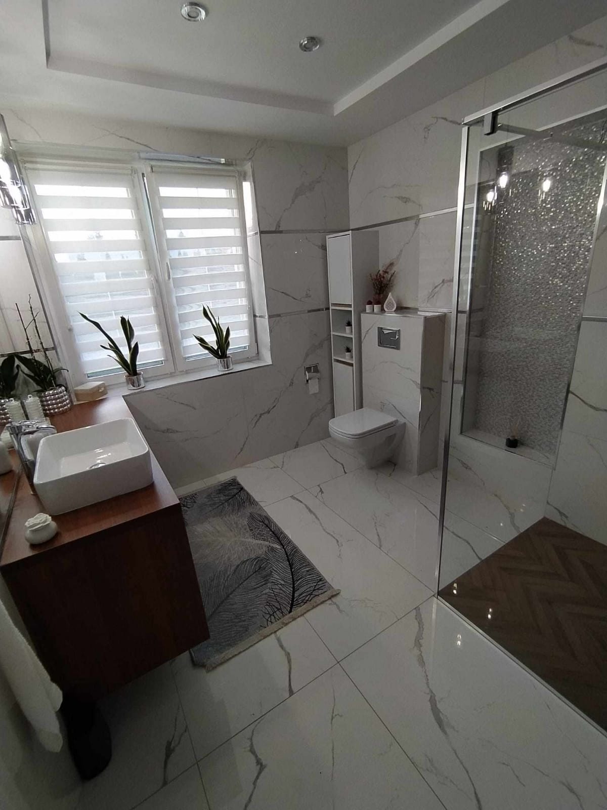 Elegancka łazienka w bieli i brązie z marmurowymi płytkami, kabiną prysznicową typu walk-in z dekoracyjną ścianą, drewnianym akcentem pod prysznicem, wiszącą toaletą i umywalką nablatową.