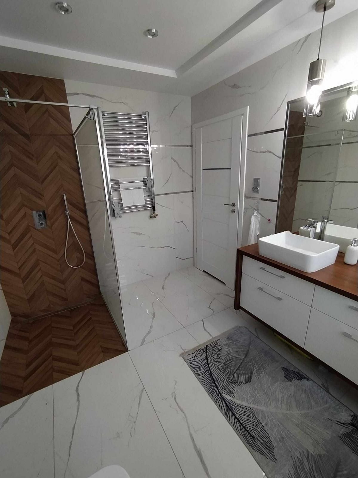 Nowoczesna łazienka z kabiną prysznicową wyłożoną drewnopodobną mozaiką w jodełkę, marmurowe płytki na ścianach i podłodze, biała umywalka nablatowa na szafce z drewnianym blatem, grzejnik...