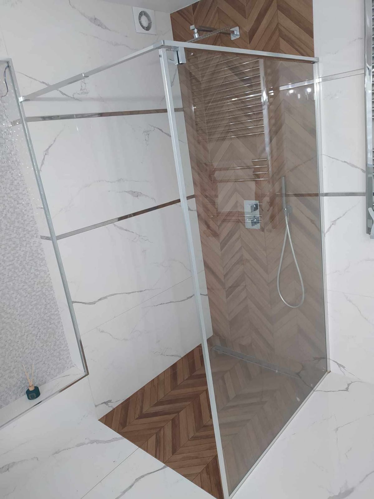 Nowoczesna kabina prysznicowa z chromowanymi profilami, drewnopodobne płytki ułożone w jodełkę na ścianie i podłodze, białe marmurowe płytki z metalicznymi listwami, widoczny panel sterowania...