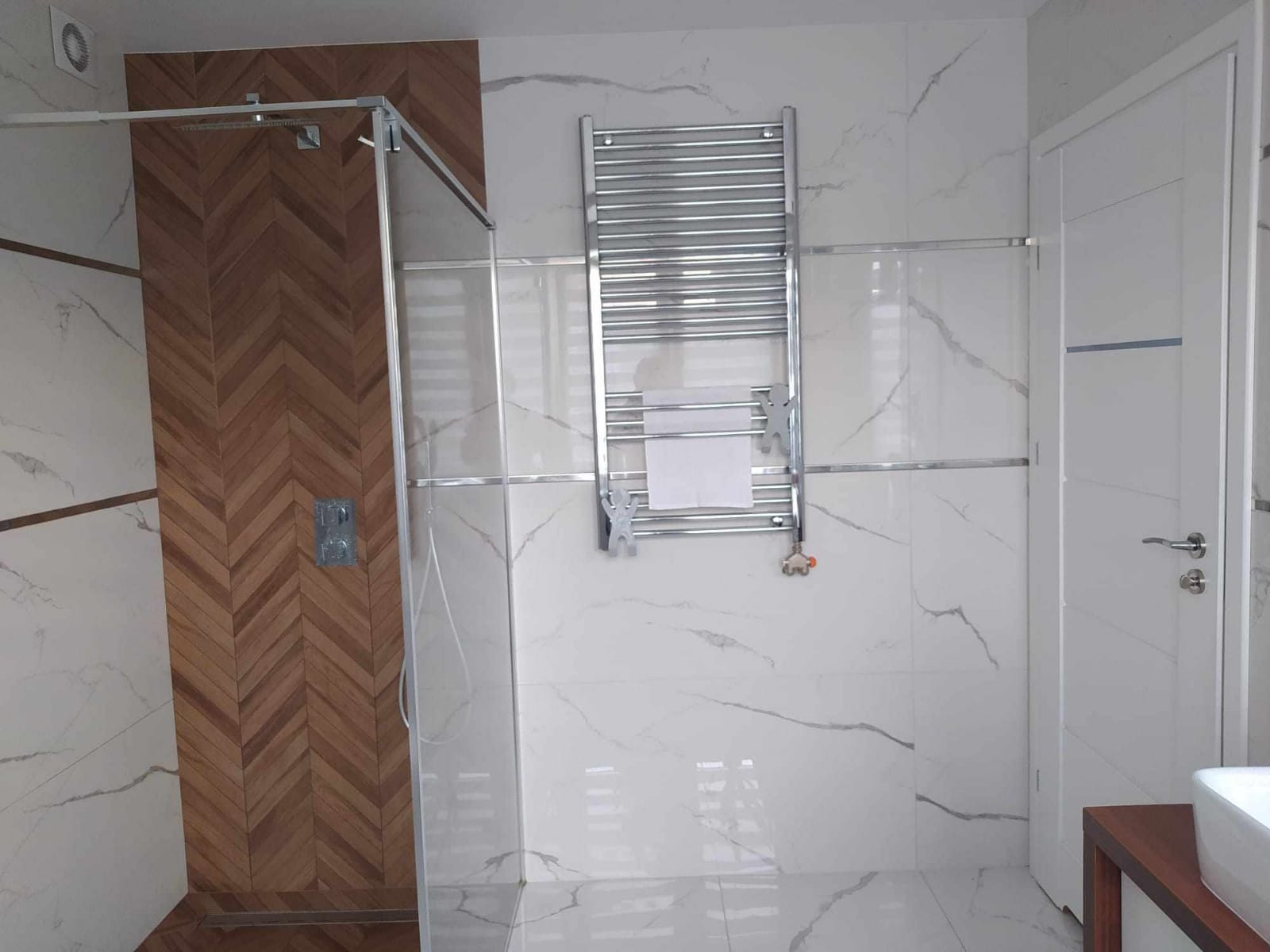 Nowoczesna łazienka z kabiną prysznicową wyłożoną drewnopodobnymi płytkami ułożonymi w jodełkę, marmurowe płytki na ścianach, grzejnik drabinkowy z ręcznikiem i białe drzwi.