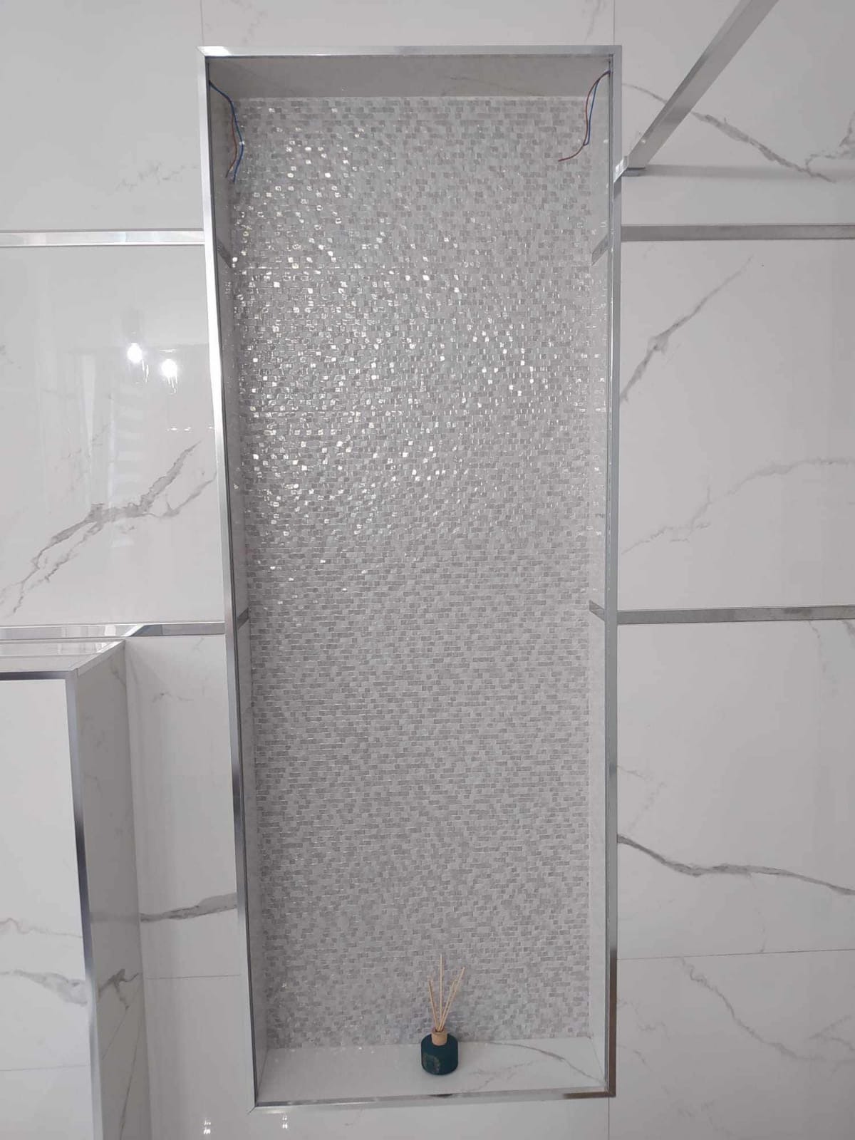 Niewielka wnęka w łazience wyłożona mozaiką w odcieniach szarości, obramowana metalowymi listwami, z dekoracyjnym dyfuzorem zapachowym na dole.