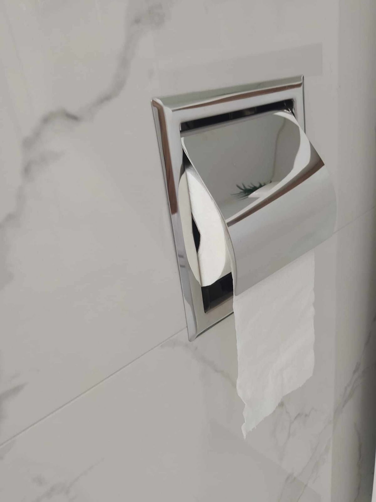 Wpuszczony w marmurową ścianę, chromowany uchwyt na papier toaletowy z częściowo rozwiniętym papierem.