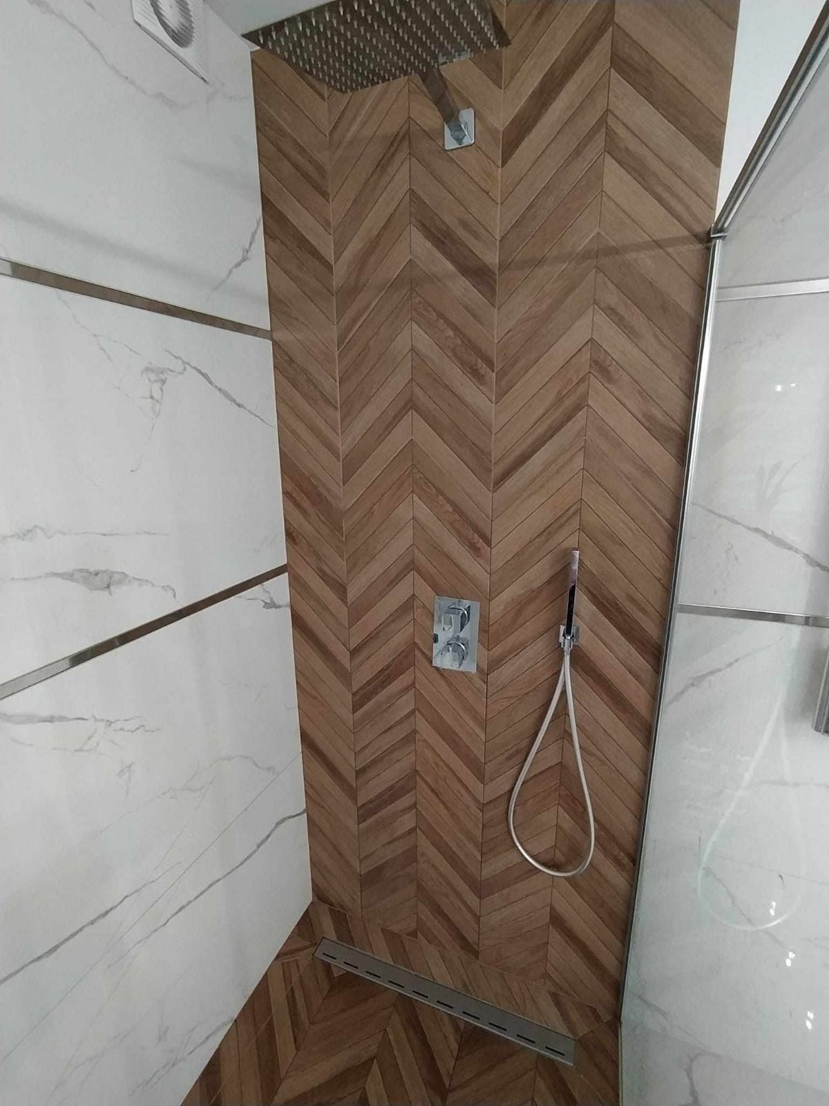 Nowoczesna kabina prysznicowa z drewnopodobnymi płytkami ułożonymi w jodełkę, białe marmurowe płytki na sąsiedniej ścianie, deszczownica i odpływ liniowy.