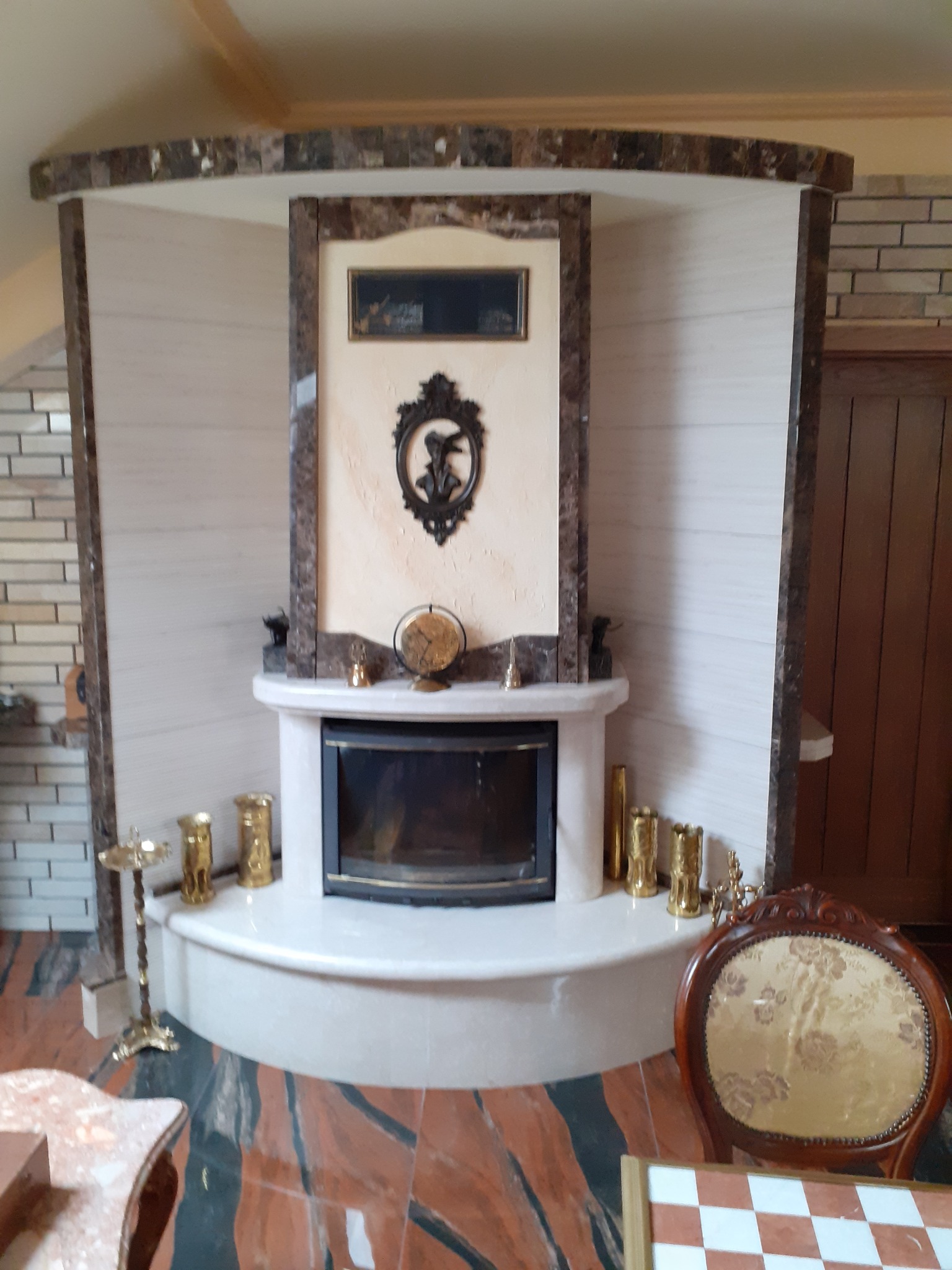 Ozdobny kominek wykończony marmurem i kamieniem dekoracyjnym, z czarnym wkładem kominkowym, otoczony złotymi świecznikami i ozdobami, na tle ceglanej ściany i drewnianych drzwi.