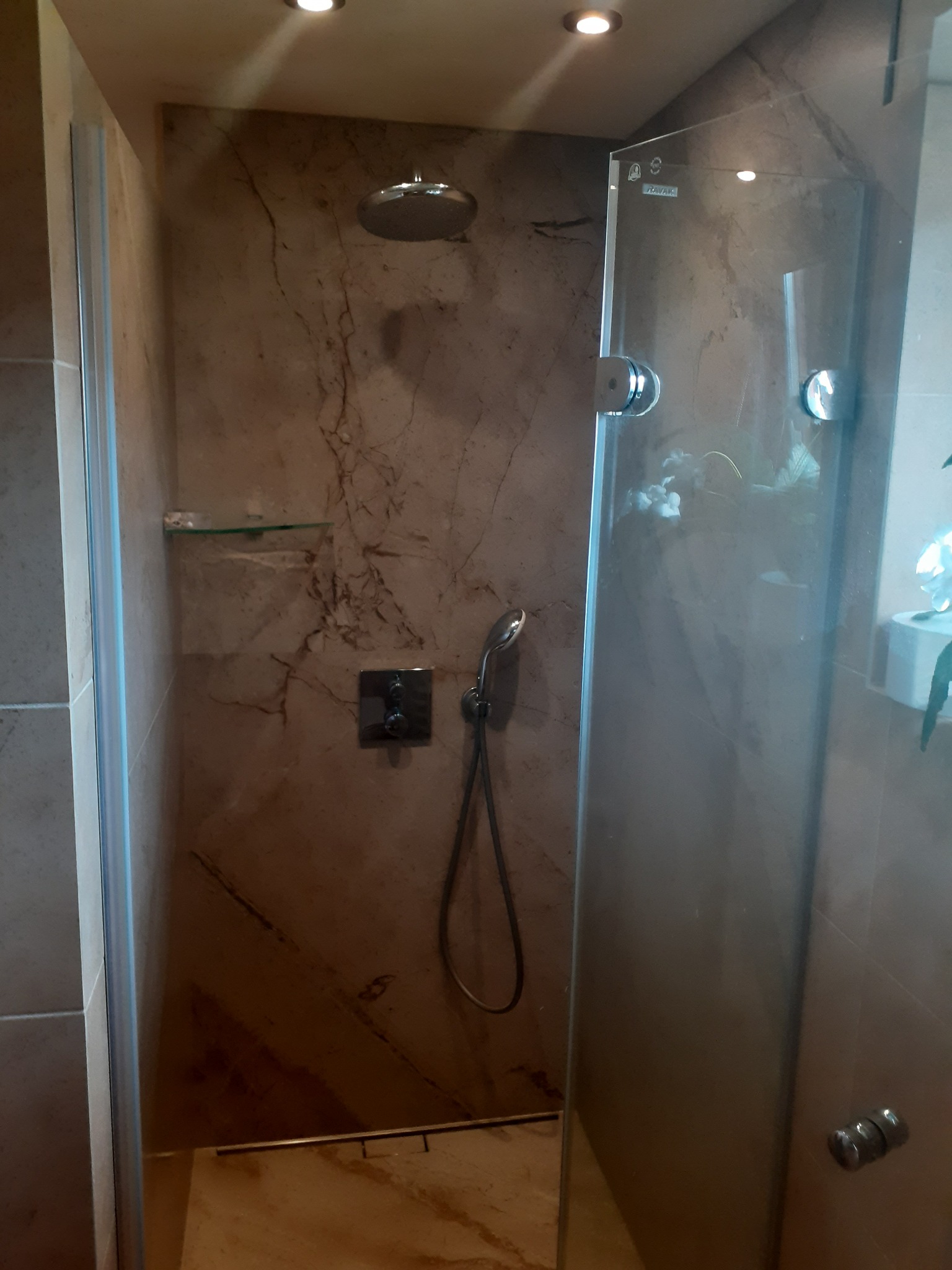 Wykończona kabina prysznicowa z przeszklonymi drzwiami, marmurowymi płytkami ściennymi w odcieniach beżu i brązu, deszczownicą sufitową i chromowaną baterią natryskową.