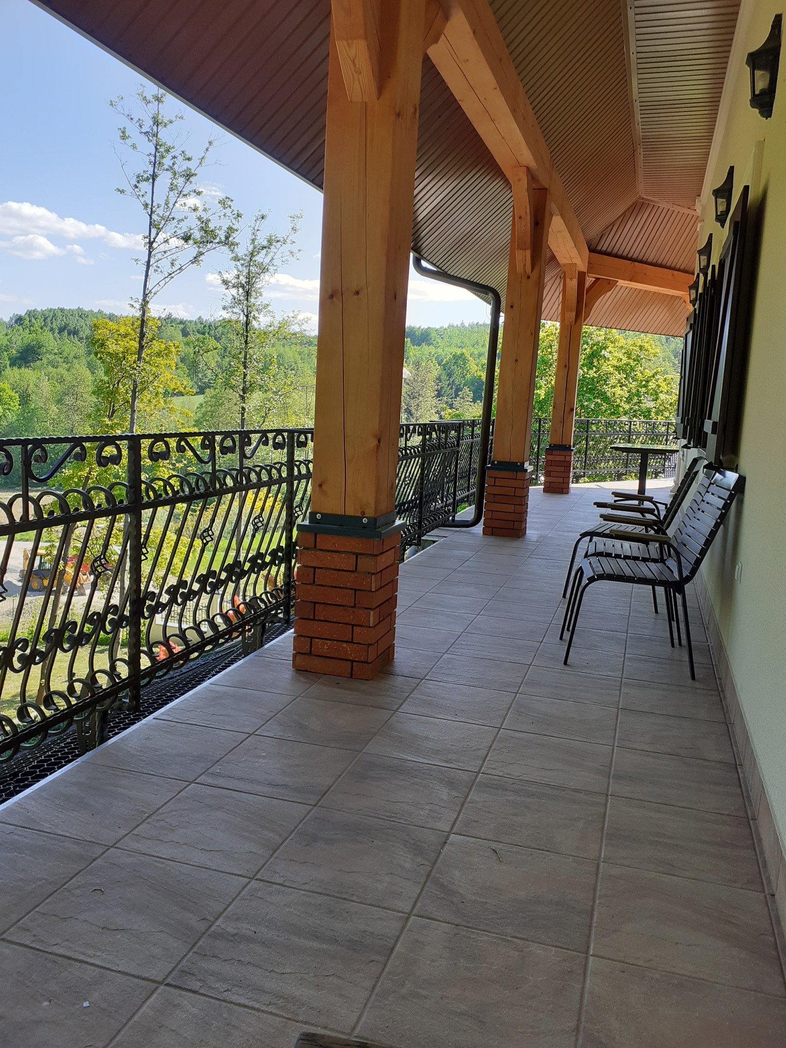 Zadaszony balkon z drewnianymi filarami na ceglanej podstawie, ozdobną metalową balustradą i szarymi płytkami podłogowymi, z widokiem na zielony krajobraz w tle.