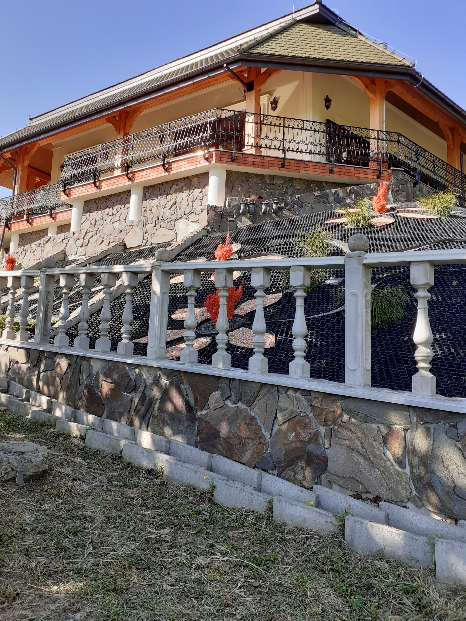 Dwupiętrowy dom z kamienną elewacją, balkonem z ozdobną balustradą i tarasem z betonową barierką, widok z dołu na tle błękitnego nieba.