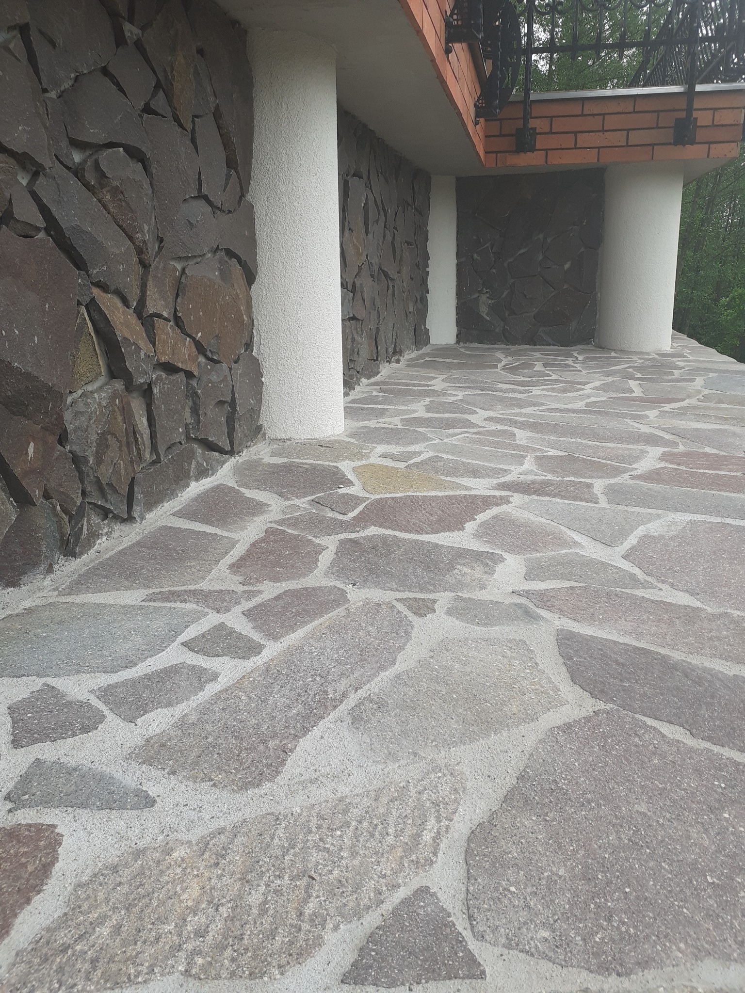 Kamienna elewacja budynku z nieregularną okładziną i kolumnami, widoczna kamienna posadzka balkonu z szarych płyt o nieregularnych kształtach.