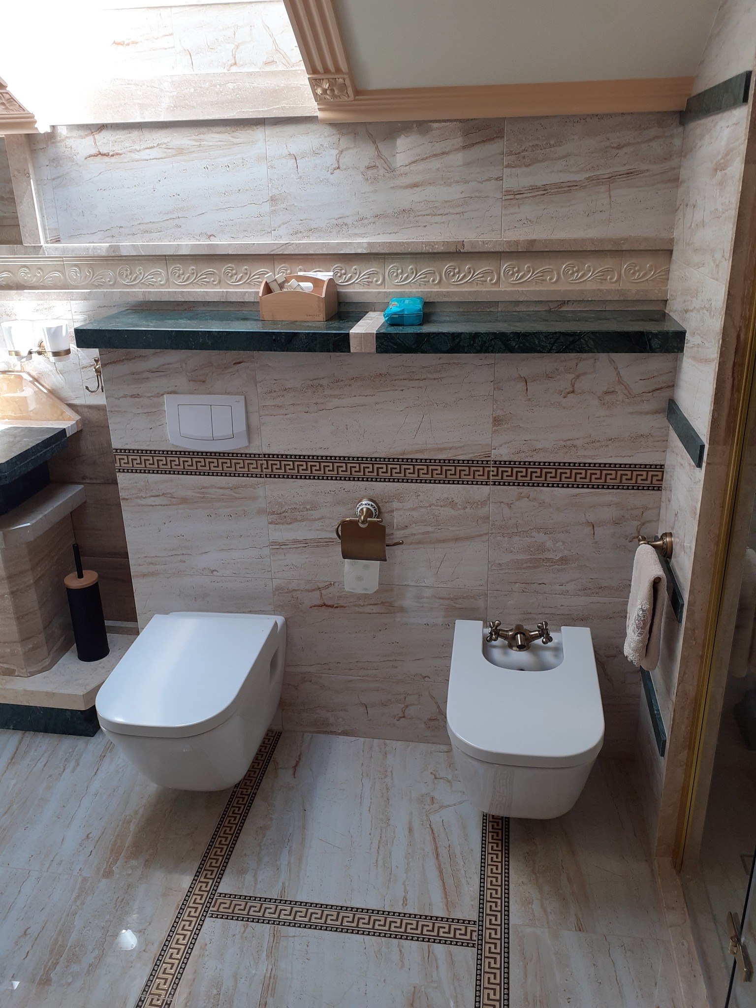 Elegancka łazienka z marmurowymi płytkami, wiszącą toaletą i bidetem, dekoracyjną listwą z motywem greckim oraz akcesoriami w kolorze złota.