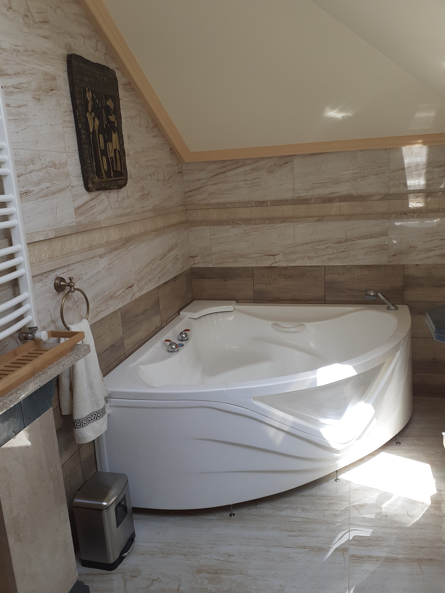 Narożna wanna z hydromasażem w łazience z marmurowymi płytkami, dekoracyjna płaskorzeźba na ścianie, widoczny kosz na śmieci i grzejnik drabinkowy.