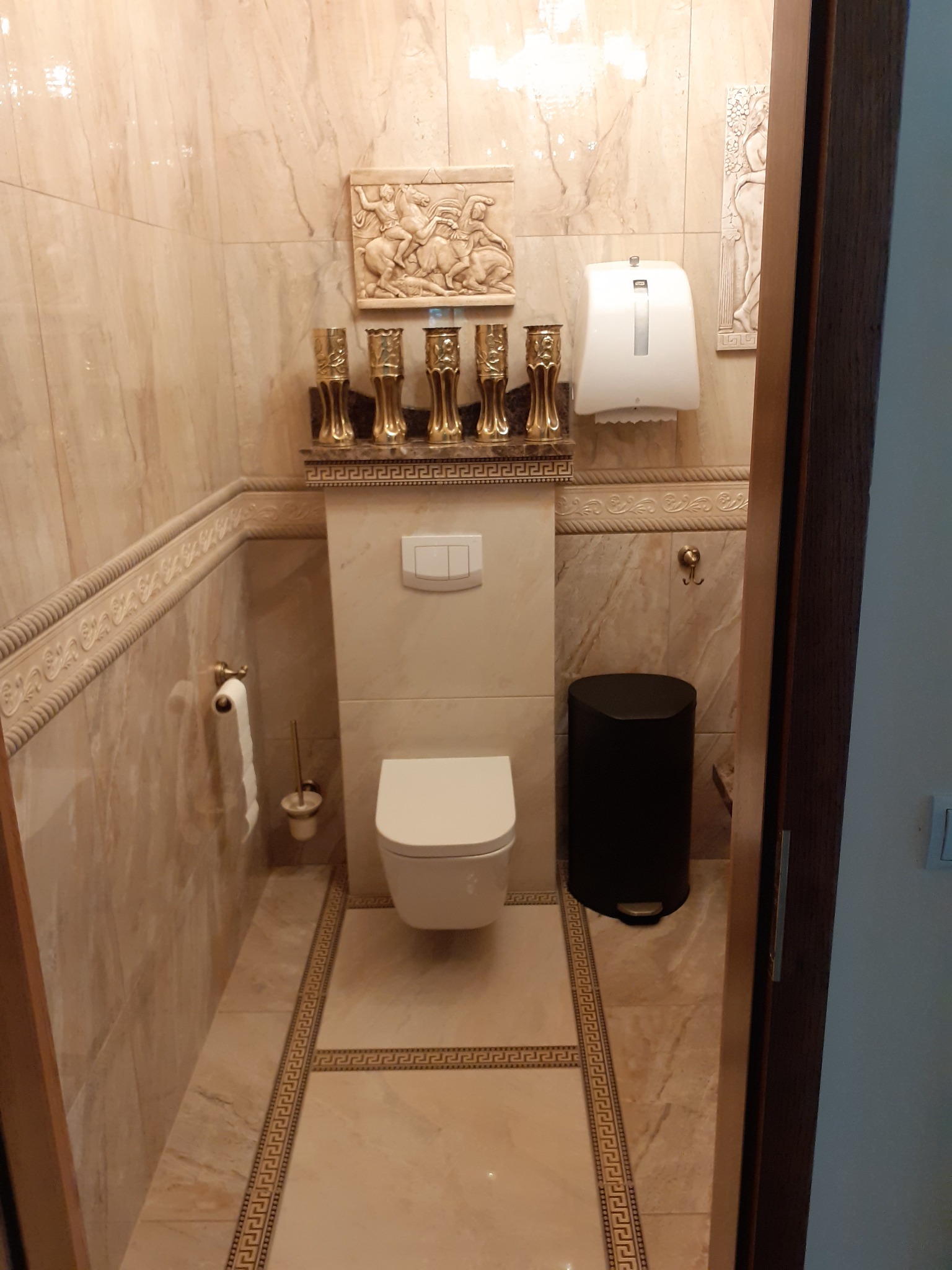Elegancka łazienka w beżowych marmurkowych płytkach z dekoracyjnymi elementami: płaskorzeźby nad toaletą i złotymi świecznikami, czarny kosz na śmieci, widok zza otwartych drzwi.
