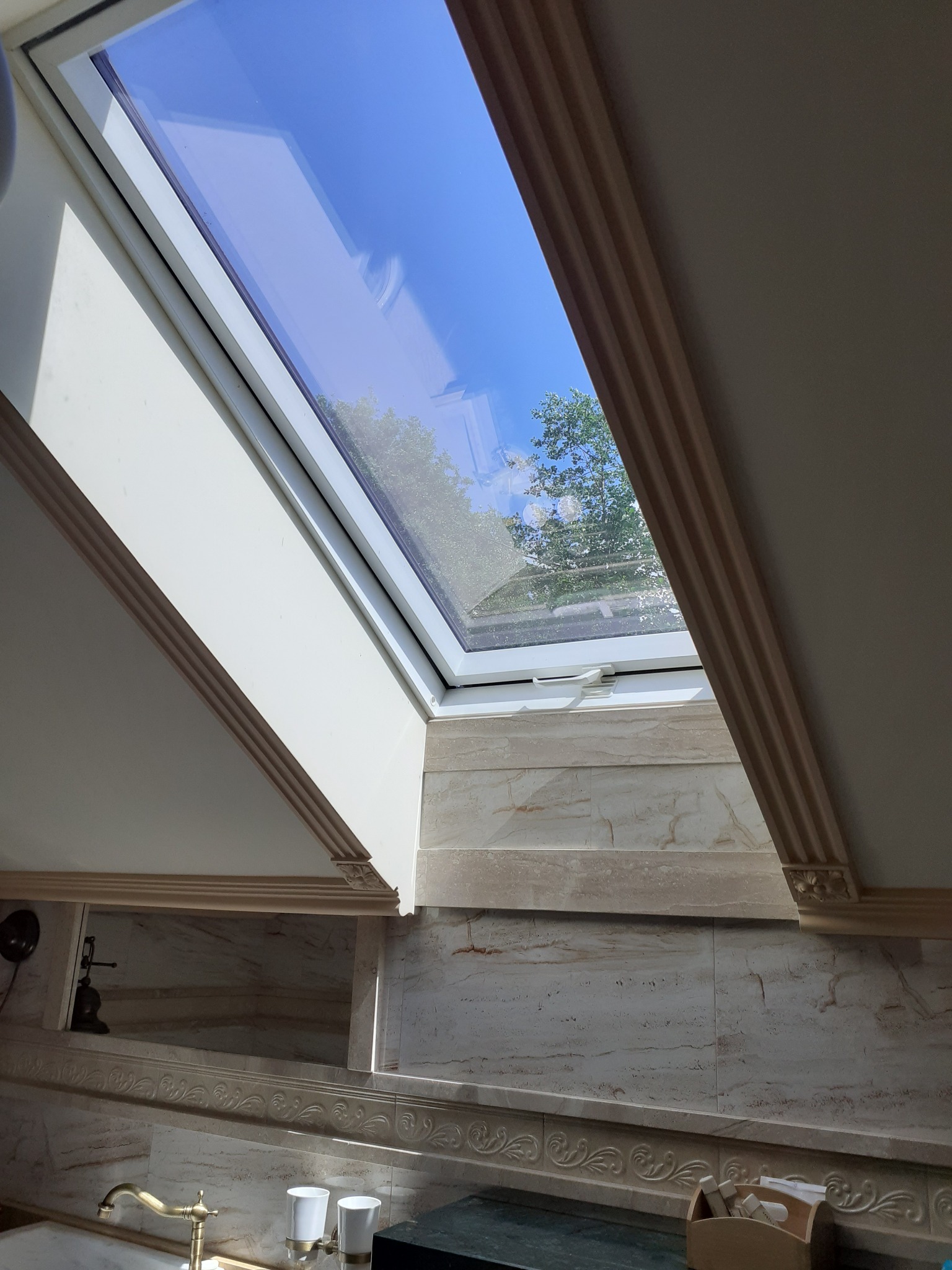 Łazienka z oknem dachowym, widok na błękitne niebo i korony drzew, marmurowe płytki ścienne z dekoracyjnym wzorem, złota bateria umywalkowa, białe kubki na szczoteczki do zębów.