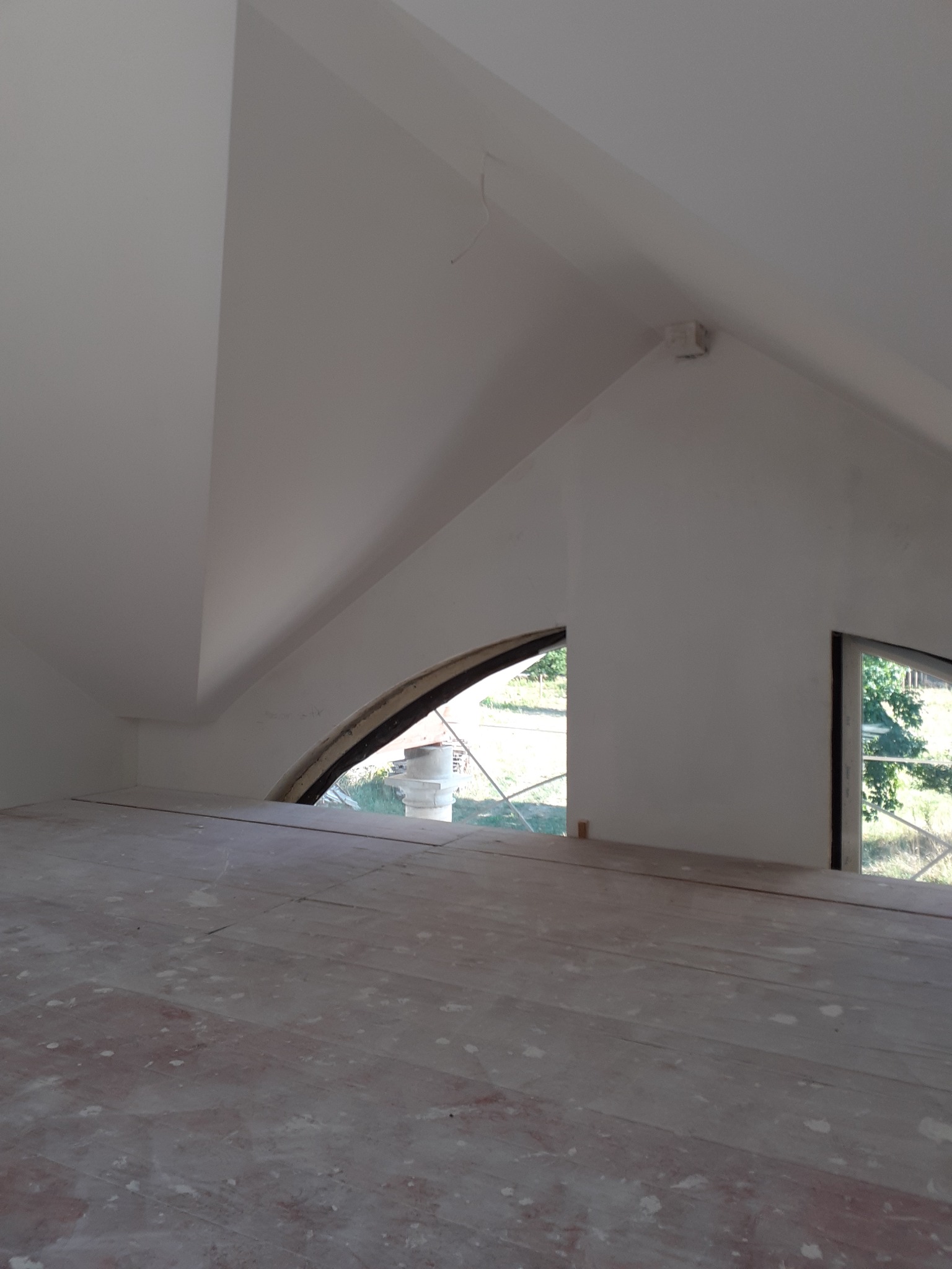 Poddasze w trakcie prac wykończeniowych, widoczne okna o nietypowych kształtach, surowe deski podłogowe pokryte pyłem, białe ściany i skosy. Widok z poziomu podłogi.