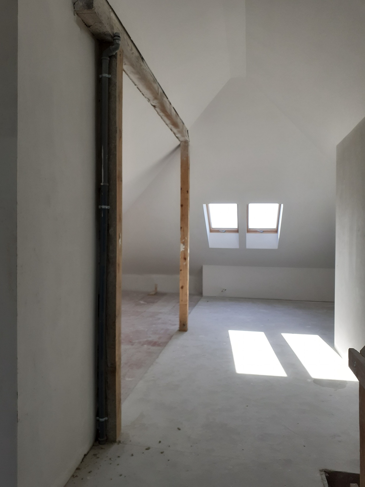 Poddasze w trakcie wykończenia, widoczne okna dachowe, surowy beton na podłodze i drewniane elementy konstrukcyjne, instalacja przy ścianie.