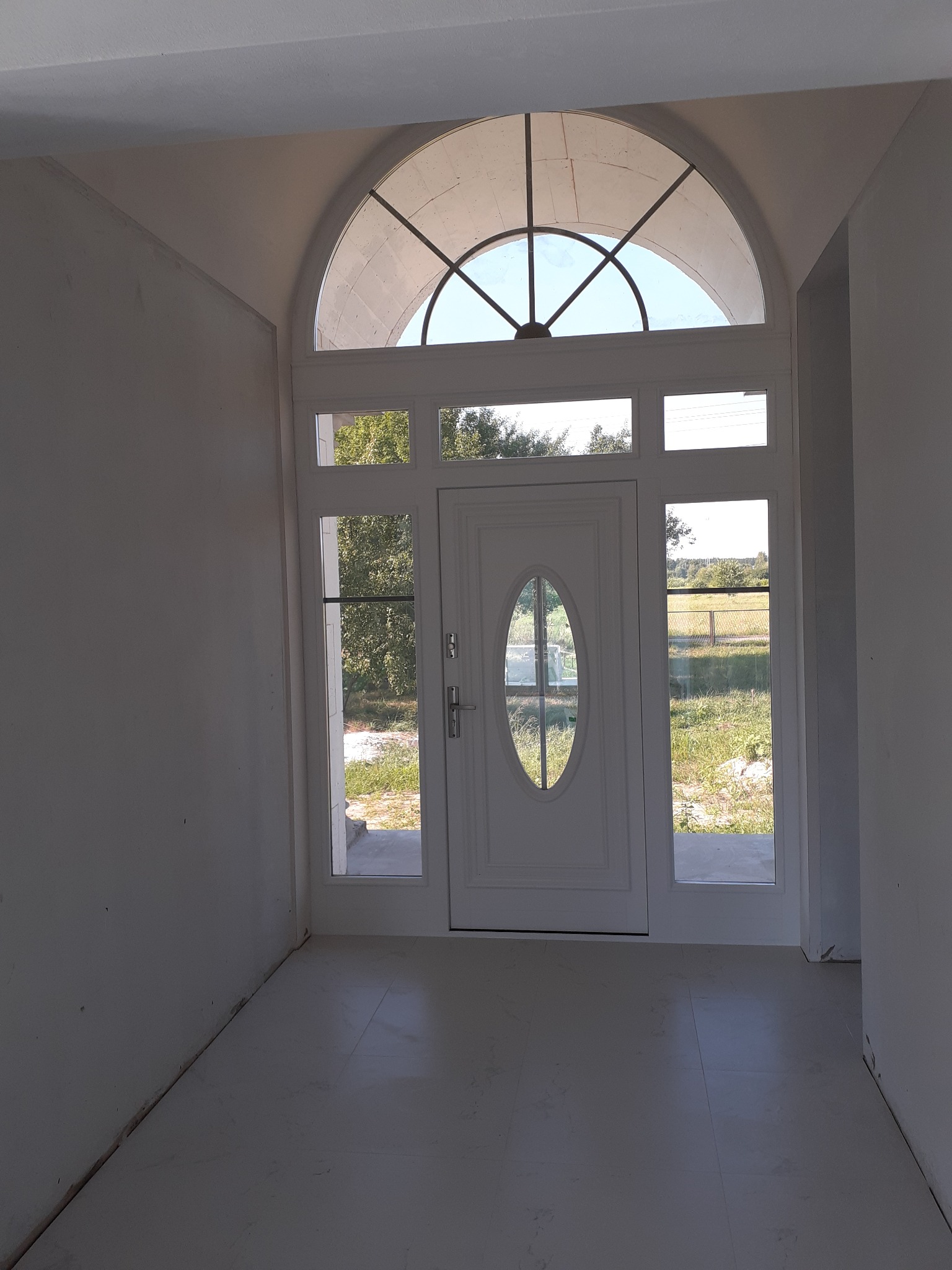 Wnętrze w trakcie wykańczania z białymi ścianami i podłogą z płytek, widok na zewnątrz przez białe drzwi z owalnym oknem i łukowym przeszkleniem nad nimi.