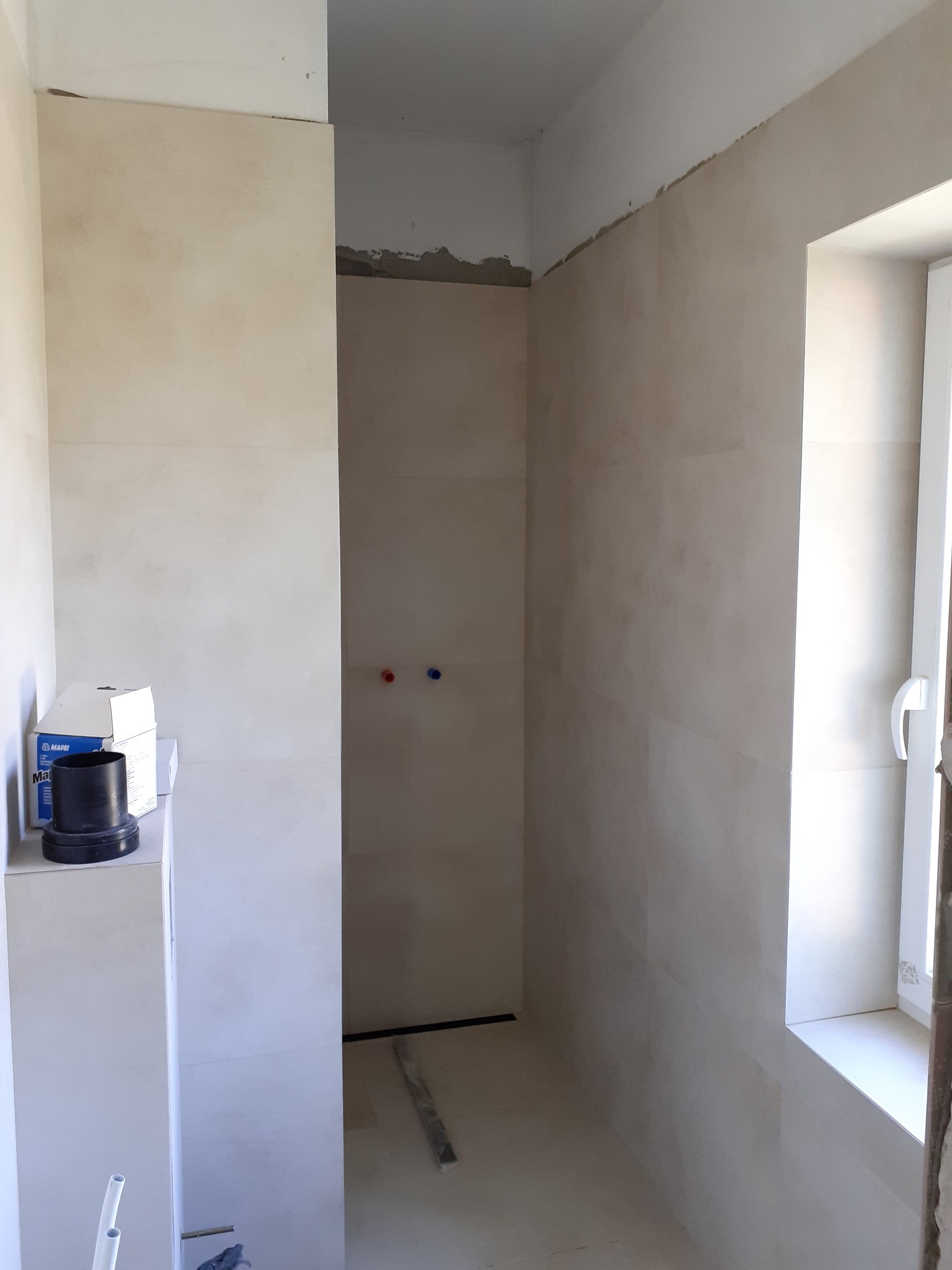 Remontowana łazienka z beżowymi płytkami na ścianach, widoczne przyłącza wodne, okno i pudełka po materiałach budowlanych na szafce.