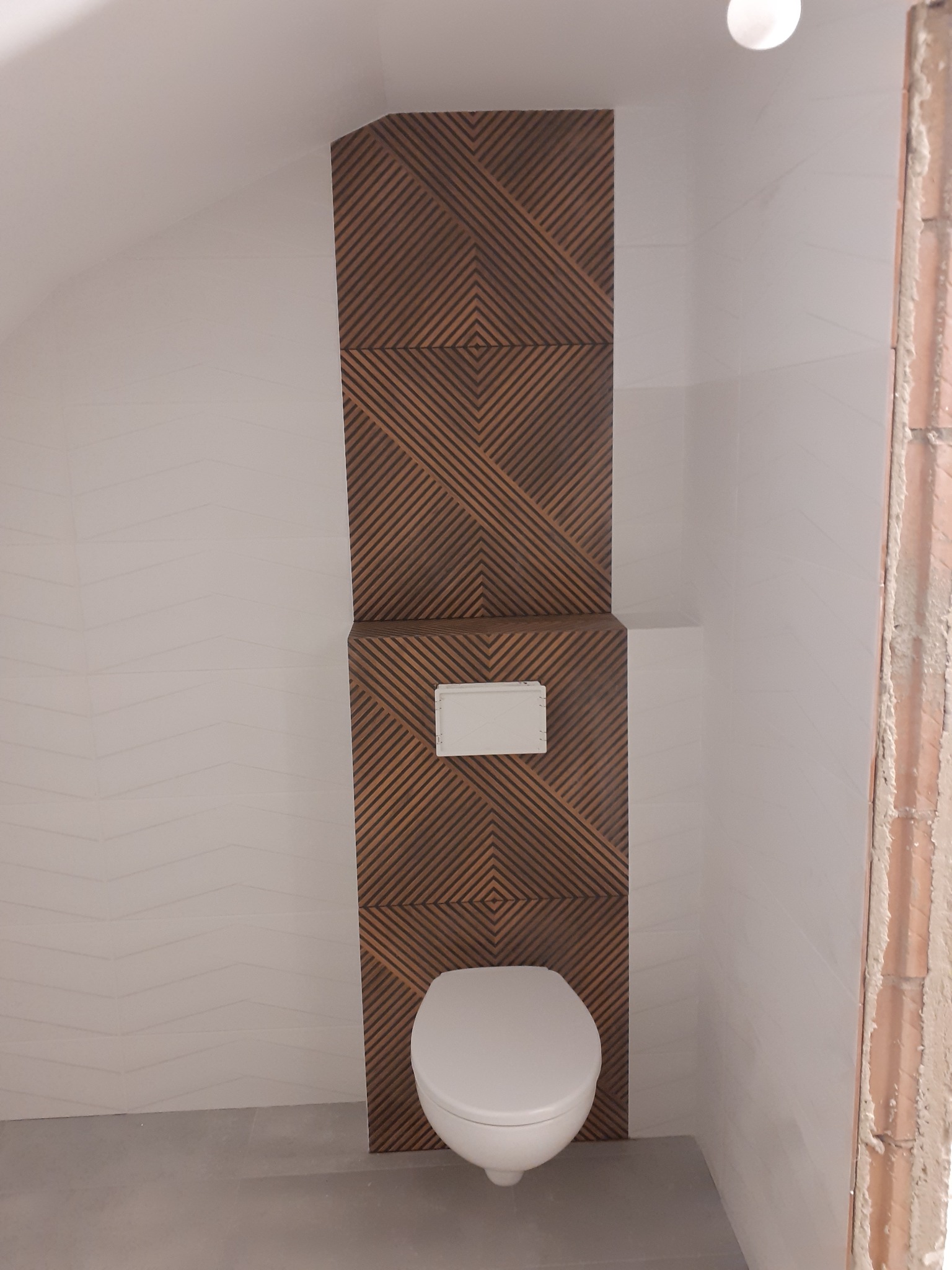 Nowoczesna toaleta podwieszana z drewnopodobną zabudową stelaża i geometrycznym wzorem, białe płytki na ścianie z delikatną falą, szara podłoga, surowy mur w narożniku.