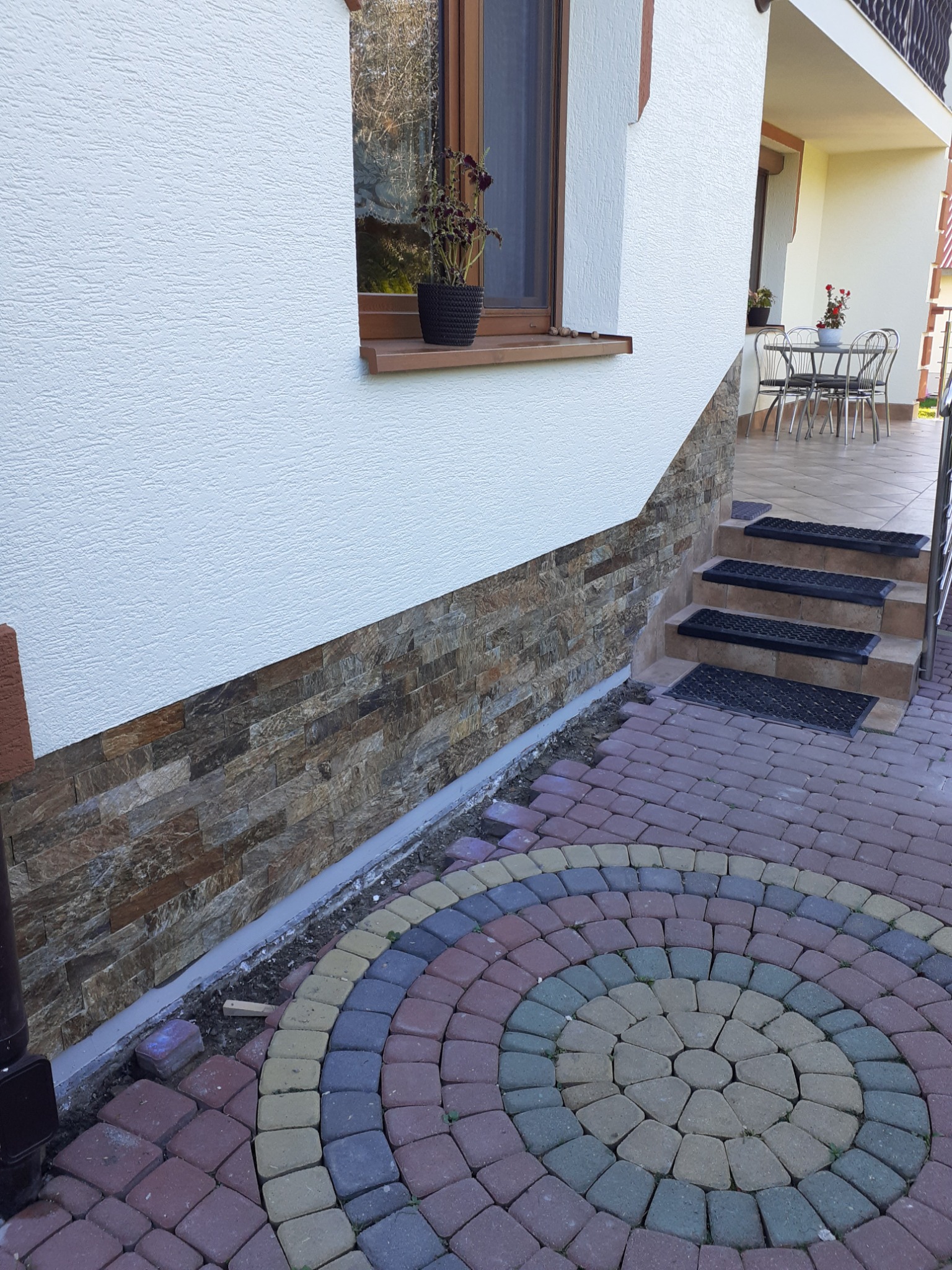 Elewacja domu z dekoracyjnym kamieniem elewacyjnym, widoczne schody z czarnymi wycieraczkami i ozdobna kostka brukowa ułożona w okrągły wzór, doniczka z kwiatami na parapecie