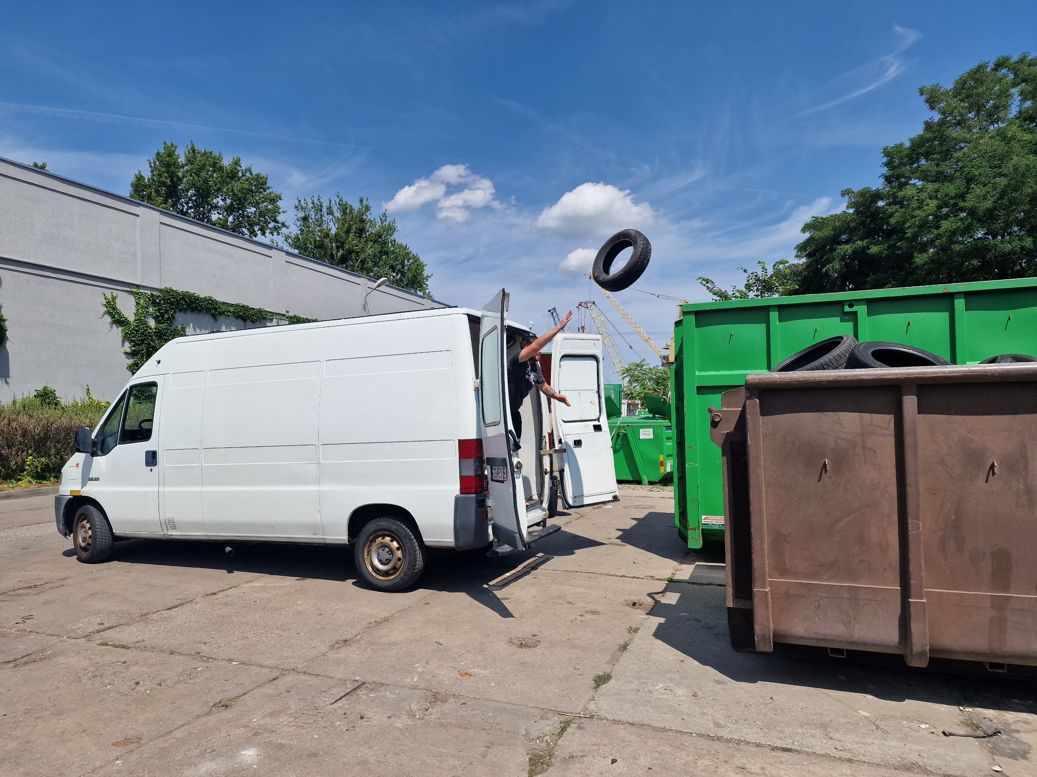 Biały furgon z otwartymi drzwiami, z którego osoba wyrzuca oponę do zielonego kontenera na tle błękitnego nieba z chmurami.