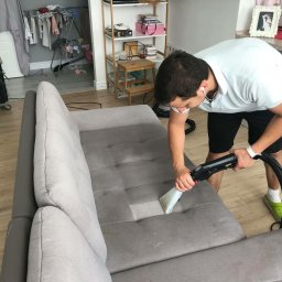 Eco Imperial Cleaning - Mężczyzna w krótkich spodenkach i zielonych klapkach czyści szarą kanapę odkurzaczem piorącym. Widoczny fragment wyczyszczonej powierzchni. W tle suszarka na pranie, regał i fragment kuchni.