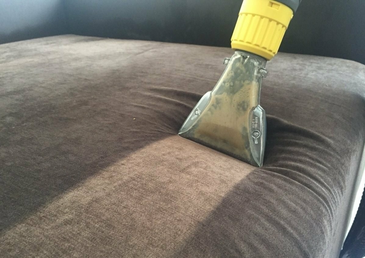 Dysza odkurzacza piorącego z widocznymi zabrudzeniami w trakcie czyszczenia brązowej, welurowej kanapy.