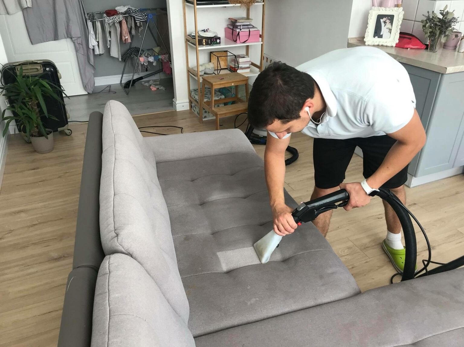Mężczyzna w krótkich spodenkach i zielonych klapkach czyści szarą kanapę odkurzaczem piorącym. Widoczny fragment wyczyszczonej powierzchni. W tle suszarka na pranie, regał i fragment kuchni.