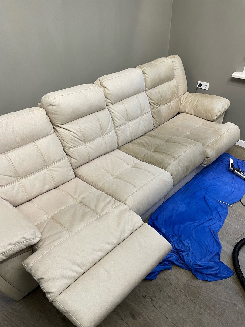 Fragment sofy z jasnej tapicerki w trakcie czyszczenia, widoczne różnice w odcieniu między wyczyszczonym a zabrudzonym fragmentem, obok niebieskie zabezpieczenie i końcówka odkurzacza piorącego.