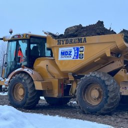 MDZ Prace ziemne - Żółta wywrotka Hydrema z ładunkiem ziemi na śniegu, widoczne logo firmy MDZ Zuterek i CAT, operator w kabinie w pomarańczowej kamizelce odblaskowej.