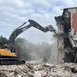 Żółto-czarna koparka gąsienicowa Volvo HB68 kruszy fragment budynku z graffiti na tle drzew i pochmurnego nieba.