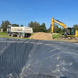 Wykop wyłożony czarną folią, obok koparka i wywrotka z ziemią, dwóch mężczyzn przy pracy, błękitne niebo w tle.