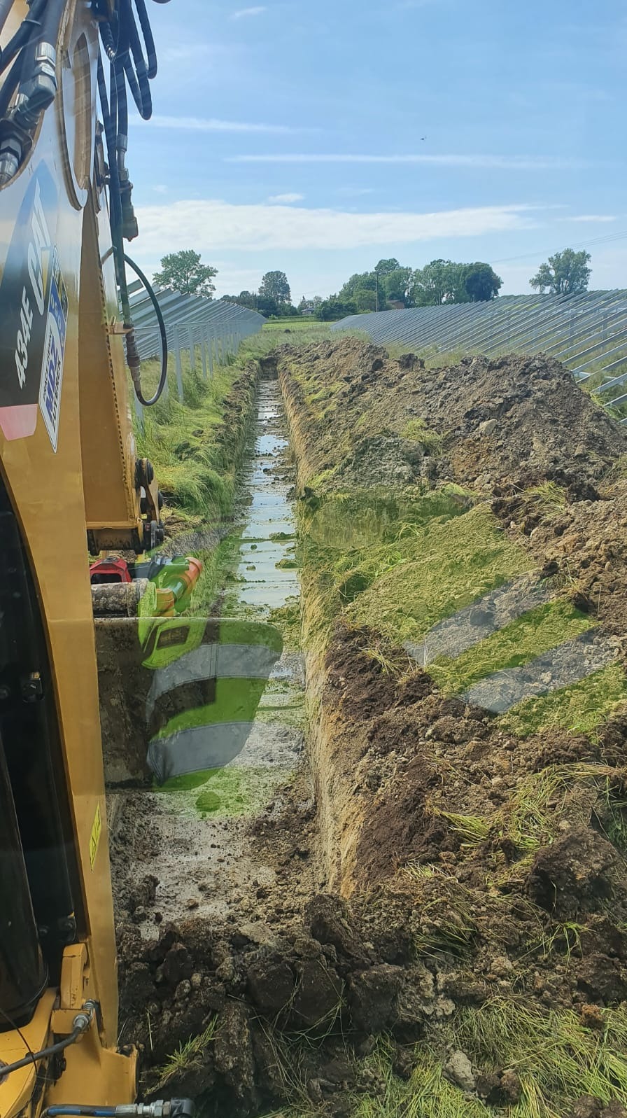 Żółta koparka gąsienicowa CAT 434E w trakcie wykonywania wąskiego wykopu w gruncie, w tle rząd paneli fotowoltaicznych i drzewa.