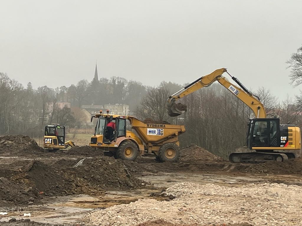 Żółta koparka CAT nabiera ziemię i wysypuje na wywrotkę Hydrema MDZ w Cerkwicy, w tle widoczny budynek z wieżą.